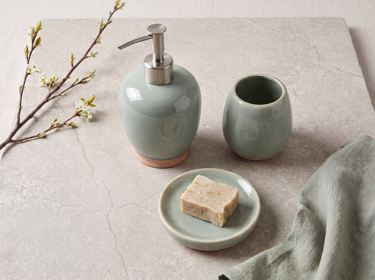 Celadon crackle-glaze ceramic soap dispenser, toothbrush holder and soap dish with a soap bar on a pale stone surface