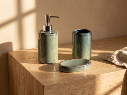 Blue-green glazed ceramic soap dispenser, toothbrush holder and oval soap dish on a sunlit honey travertine ledge.