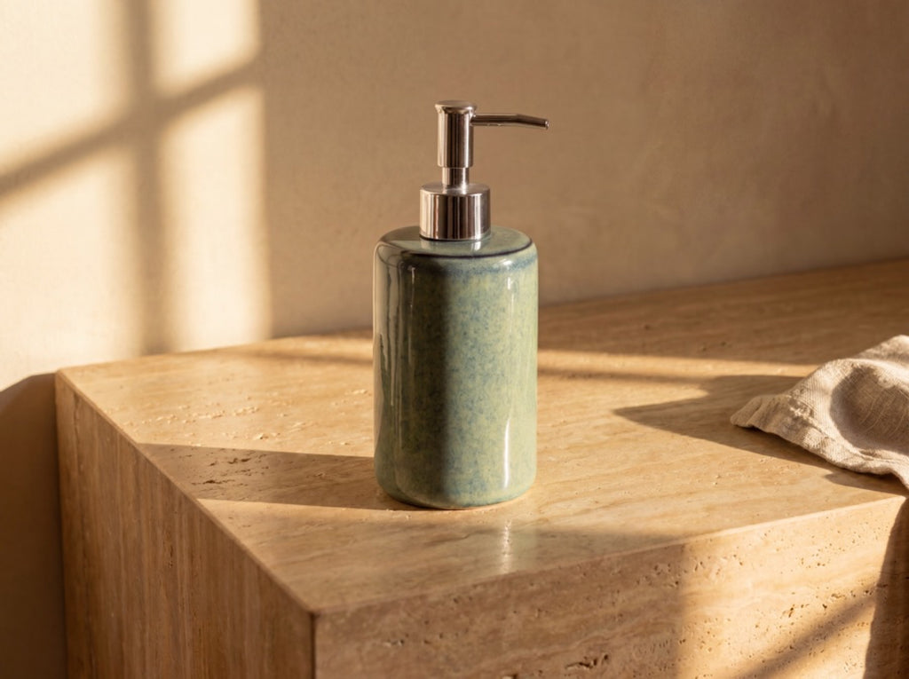 Blue-green ceramic soap dispenser with a silver pump standing alone on a warm travertine ledge in golden light.