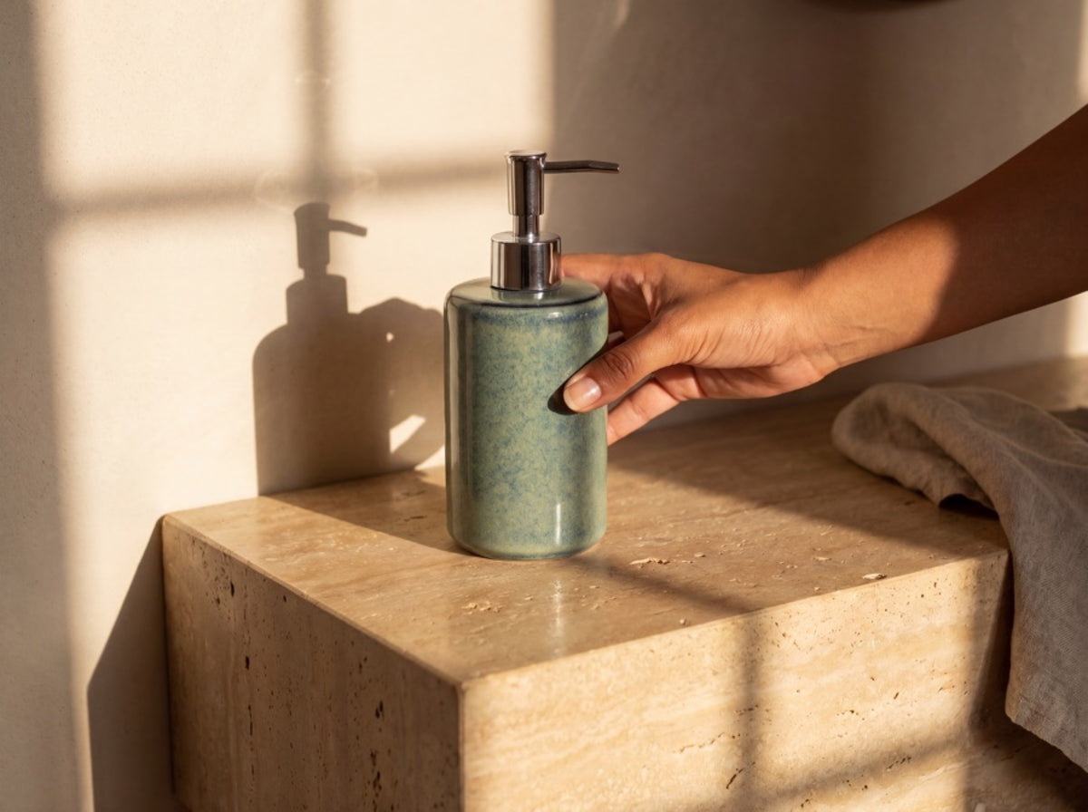 A hand resting beside the 19 cm blue-green ceramic soap dispenser on a stone counter to show its real size.