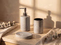 Light grey heathered ceramic soap dispenser, cylinder holder and oval soap dish on a travertine vanity in golden sunlight.