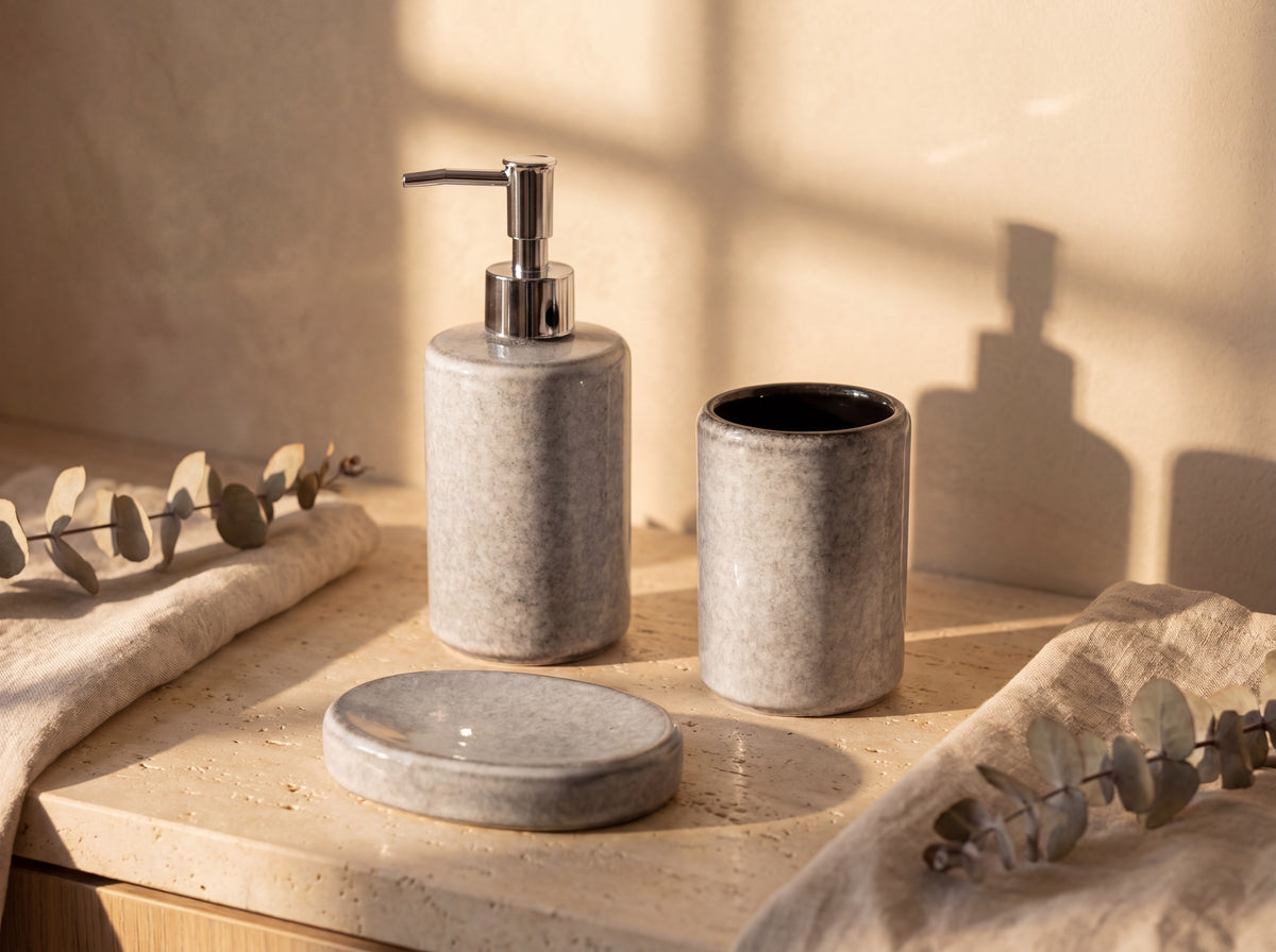 Light grey heathered ceramic soap dispenser, cylinder holder and oval soap dish on a travertine vanity in golden sunlight.