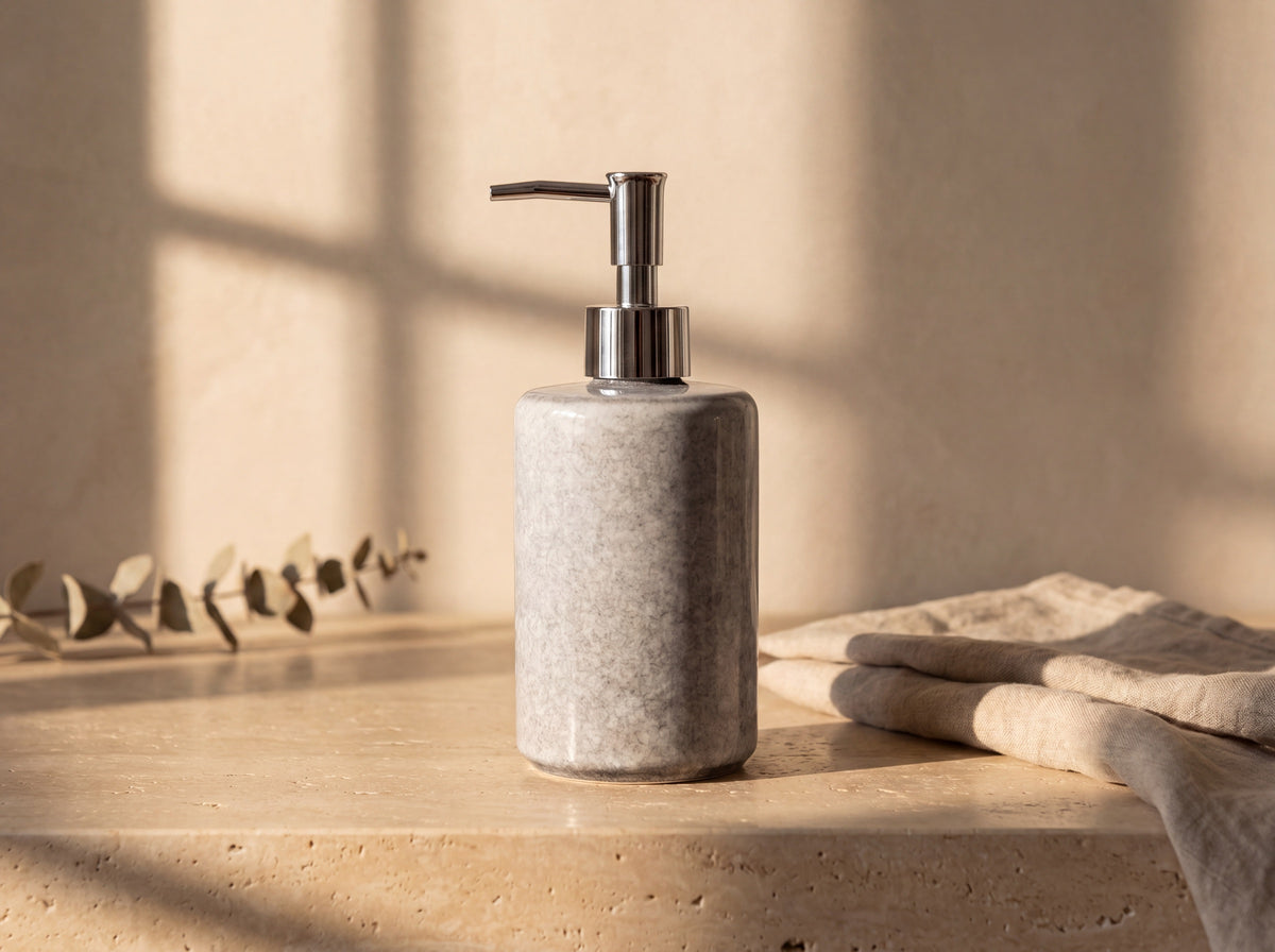Tall grey glazed ceramic soap dispenser with a chrome pump on a travertine ledge with linen and eucalyptus.