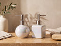 Round and square iridescent pearl ceramic soap dispensers with brushed silver pumps on a warm travertine surface against honeyed plaster, with an olive branch in a speckled cream vase and linen in golden light.