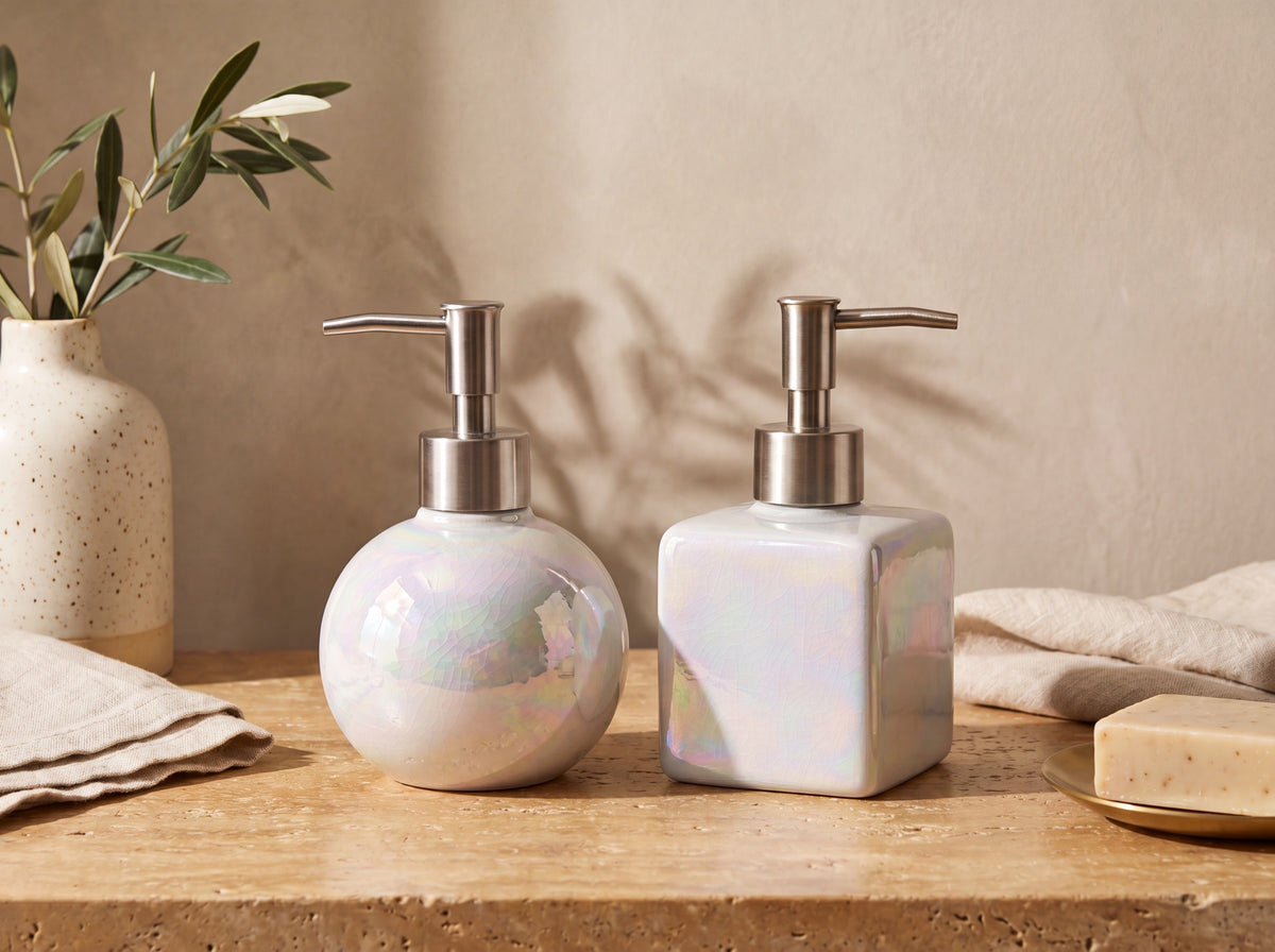 Round and square iridescent pearl ceramic soap dispensers with brushed silver pumps on a warm travertine surface against honeyed plaster, with an olive branch in a speckled cream vase and linen in golden light.