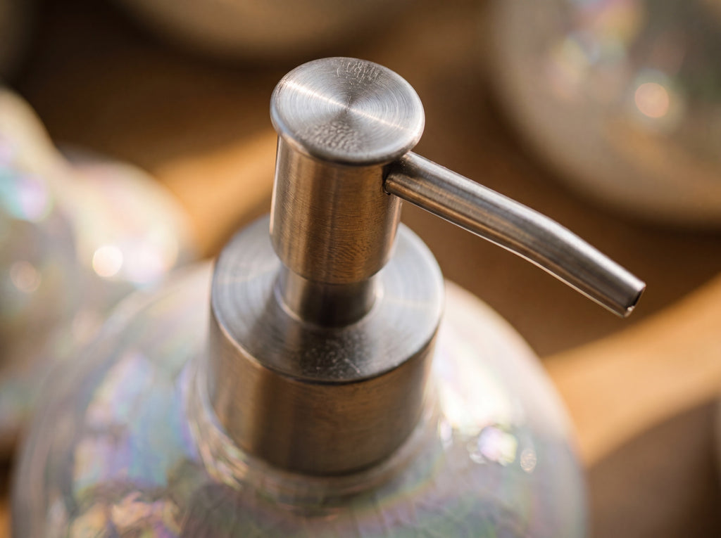 Close-up of the brushed silver stainless-steel pump head and nozzle on top of the iridescent pearl ceramic dispenser in warm golden light.