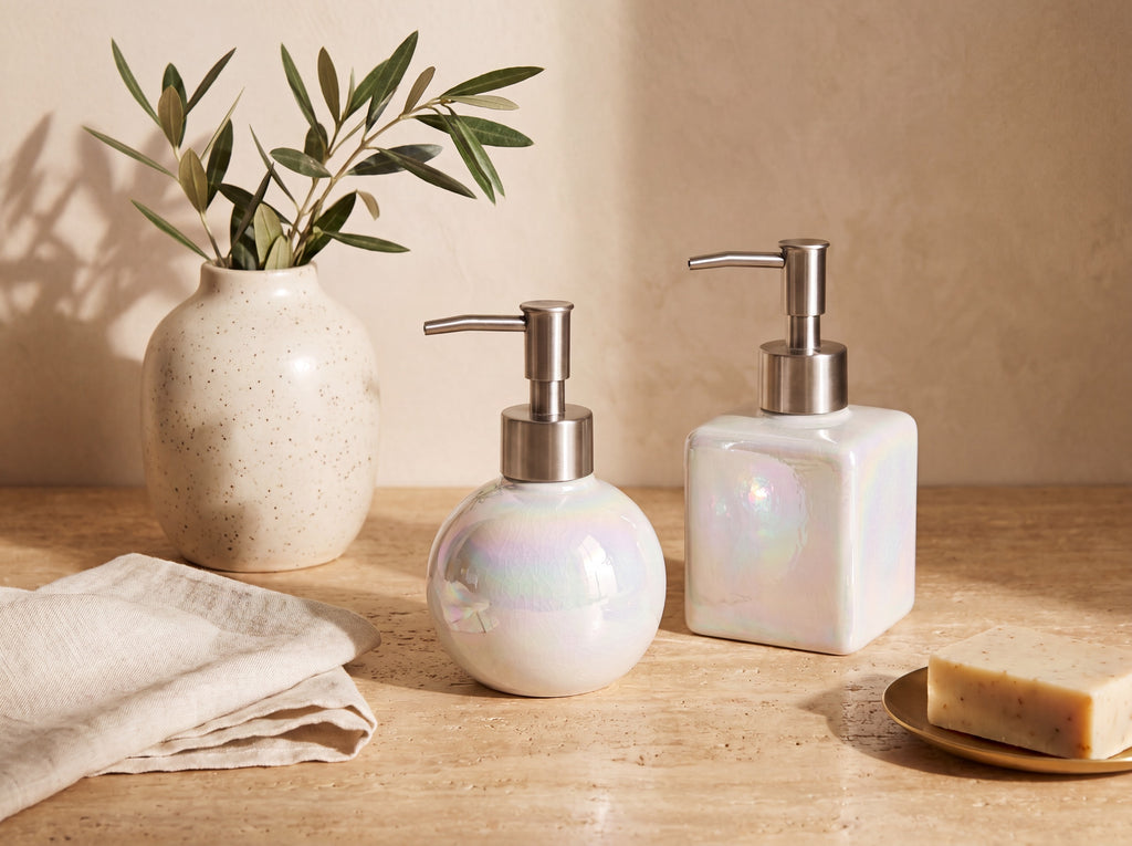 Round and square iridescent pearl ceramic dispensers styled together on warm travertine with an olive branch in a speckled vase, cream linen, and a soap bar on a brass dish.