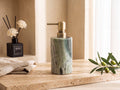 Emerald-green natural marble lotion dispenser with a brushed brass pump on a warm travertine ledge, green-and-white veining, black reed diffuser and olive stem beside it.