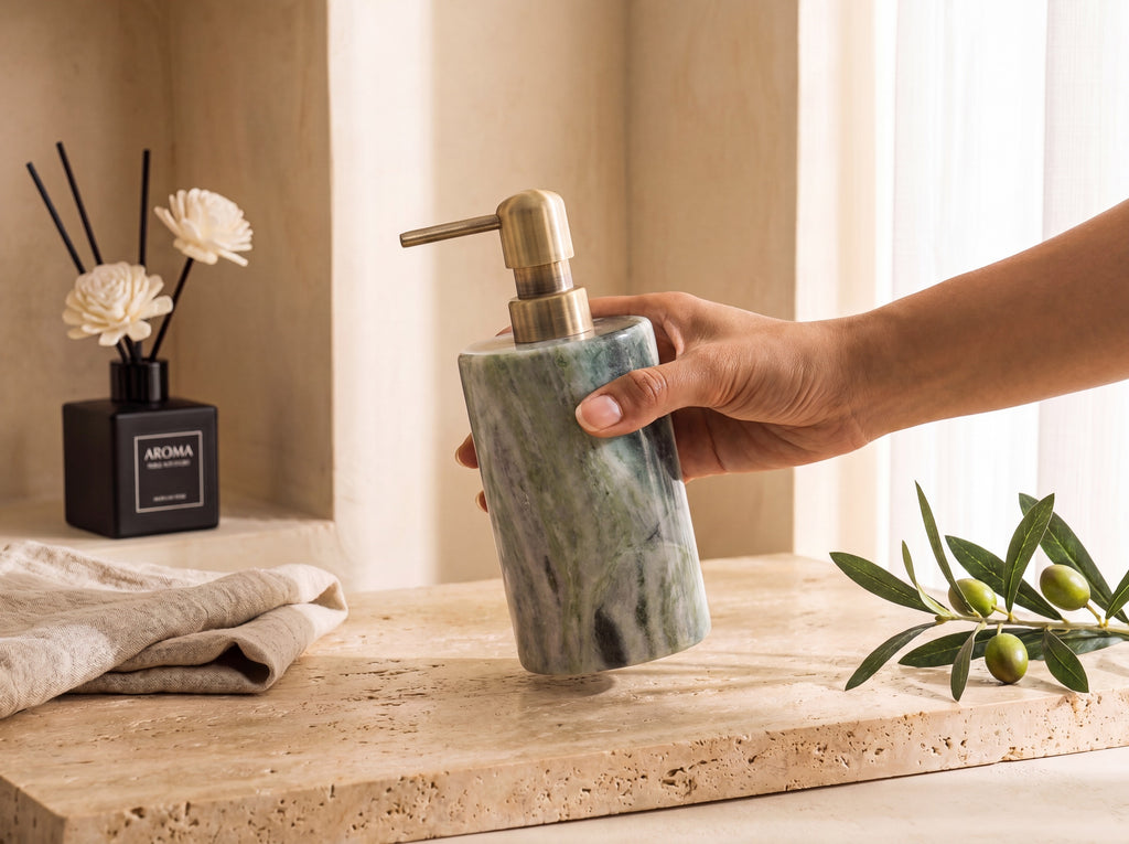 A hand with warm tan skin holding a green marble lotion dispenser to show its real-world size, warm travertine spa setting.
