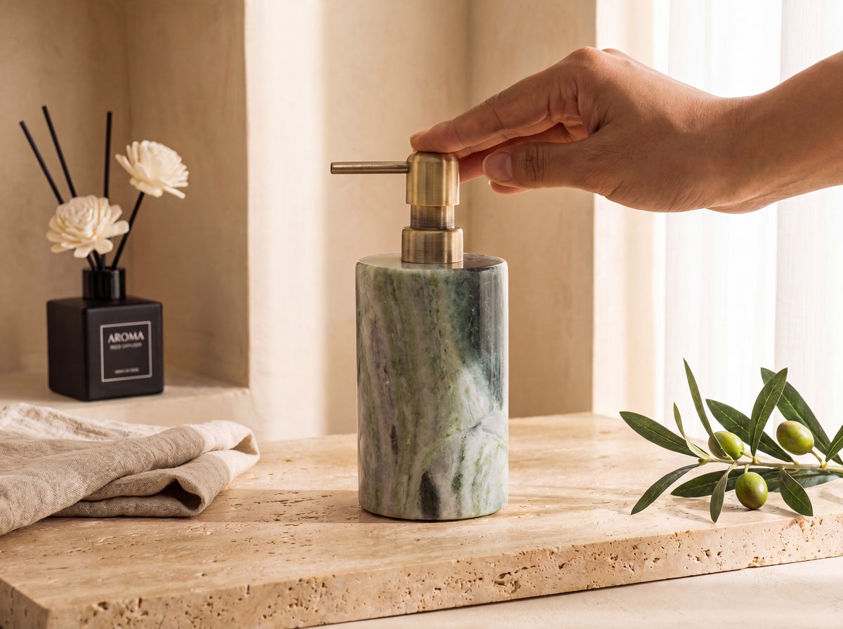 A hand pressing the brushed brass pump of a green marble lotion dispenser on a warm bathroom vanity in soft daylight.