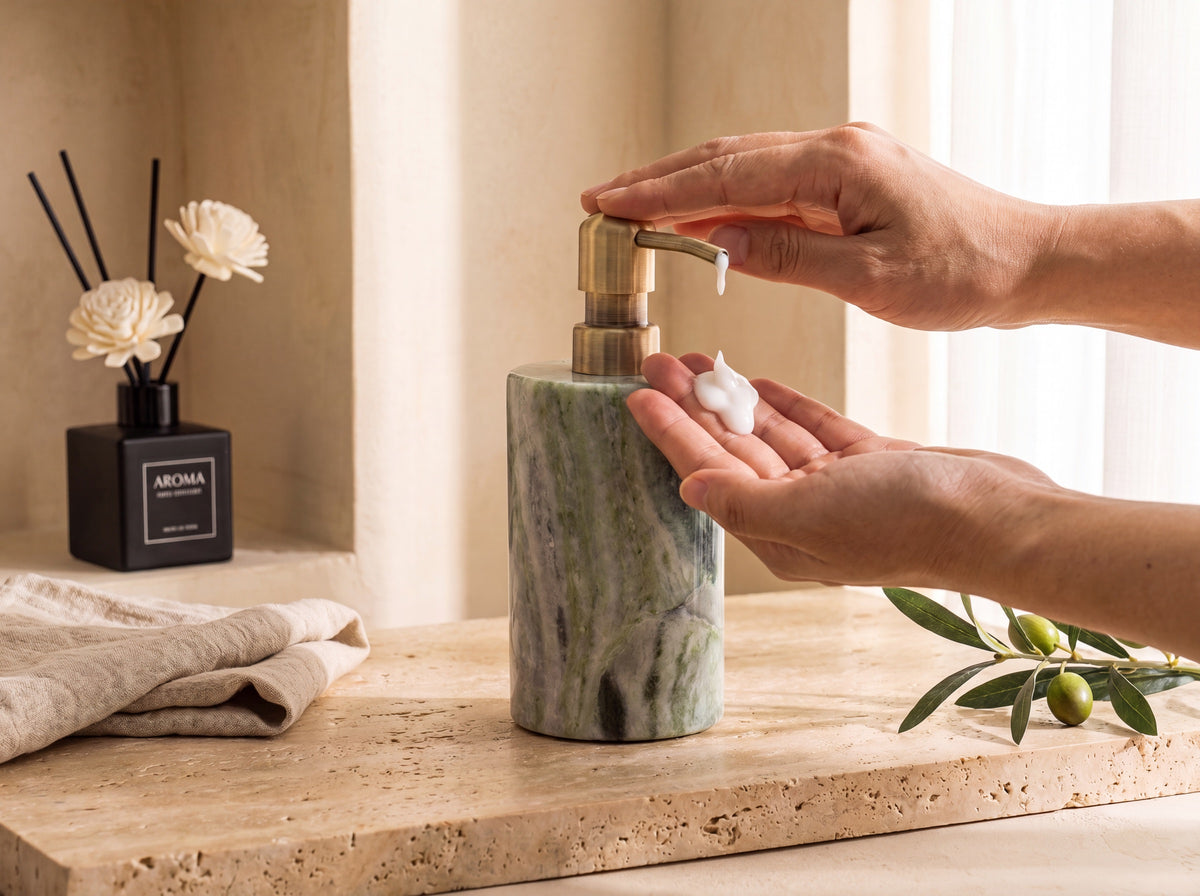A hand presses the brushed brass pump of a green marble lotion dispenser while the other palm catches a dollop of white lotion, warm travertine spa setting.