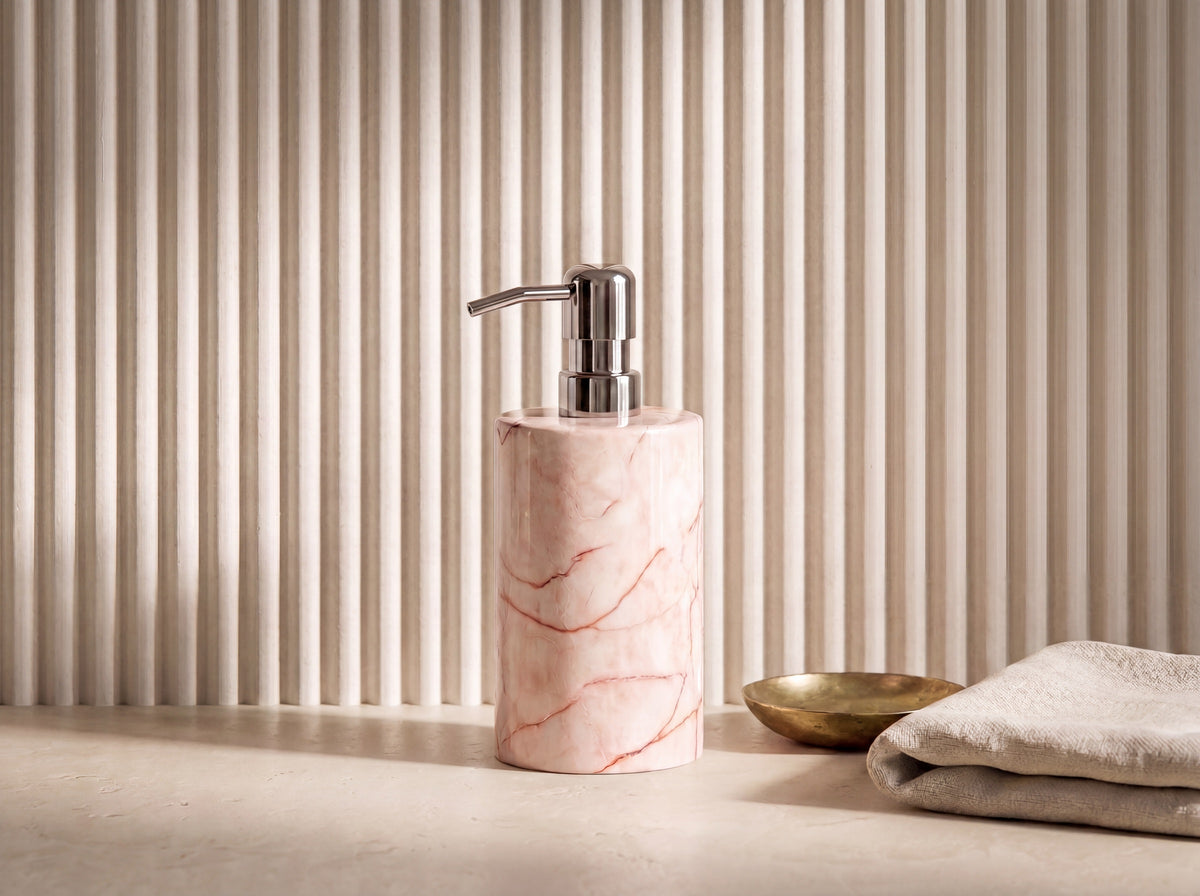 Three-quarter view of a pink marble soap dispenser showing the chrome pump spout and rose-veined stone body against a fluted cream wall.