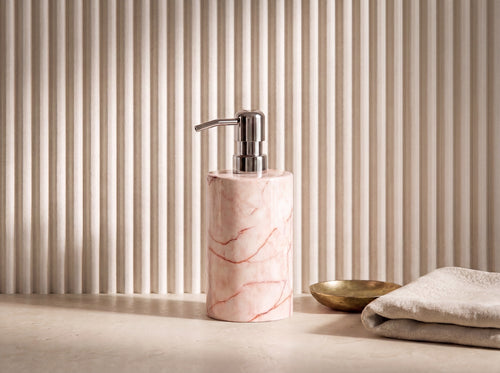 Three-quarter view of a pink marble soap dispenser showing the chrome pump spout and rose-veined stone body against a fluted cream wall.