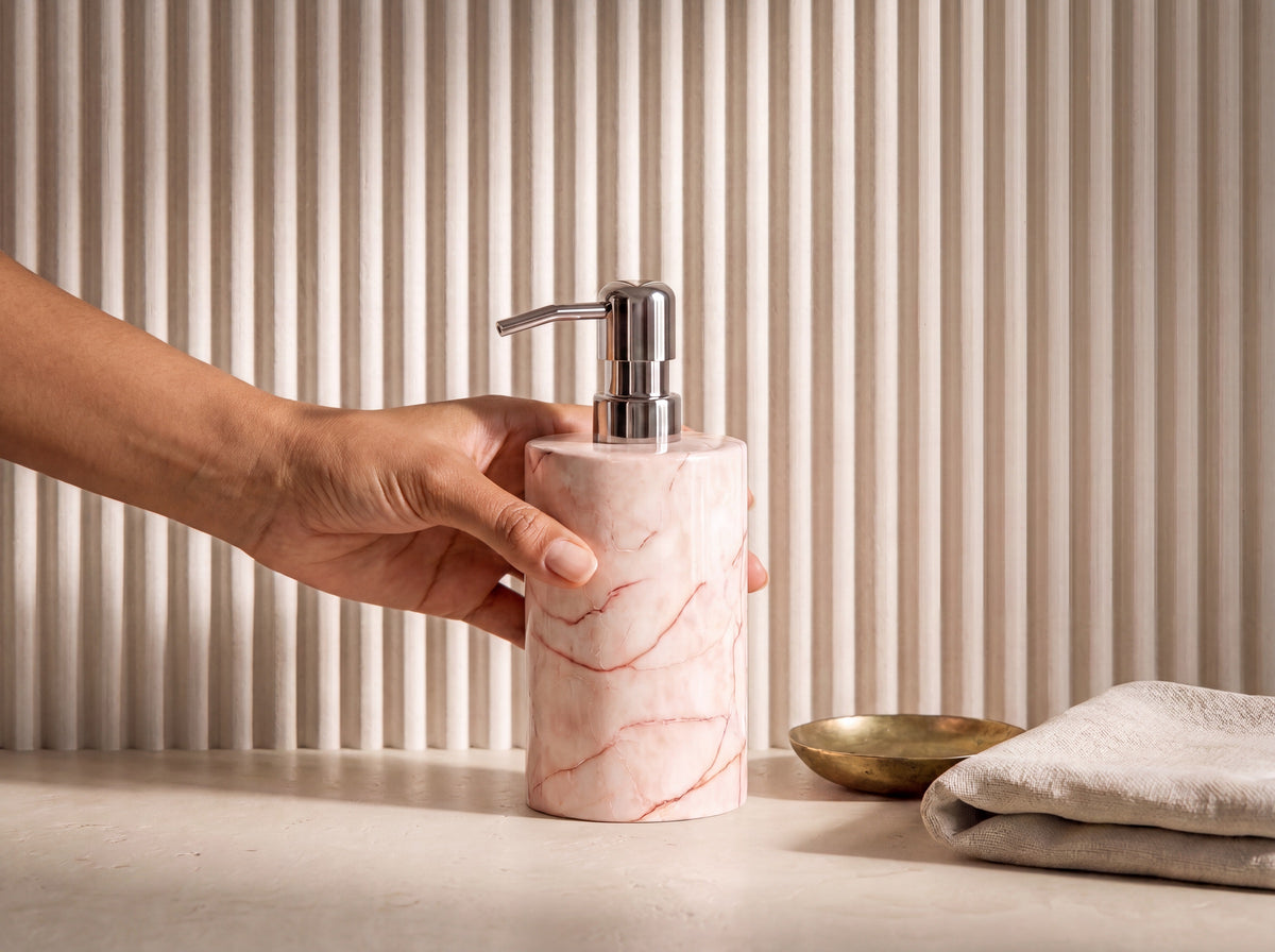 A hand with warm tan skin holding a pink marble soap dispenser to show its real-world size, bright fluted cream bathroom setting.