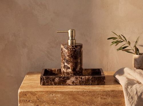 Round dark brown marble soap dispenser with an antique brass pump standing on a matching rectangular marble tray on a travertine ledge in low golden light.