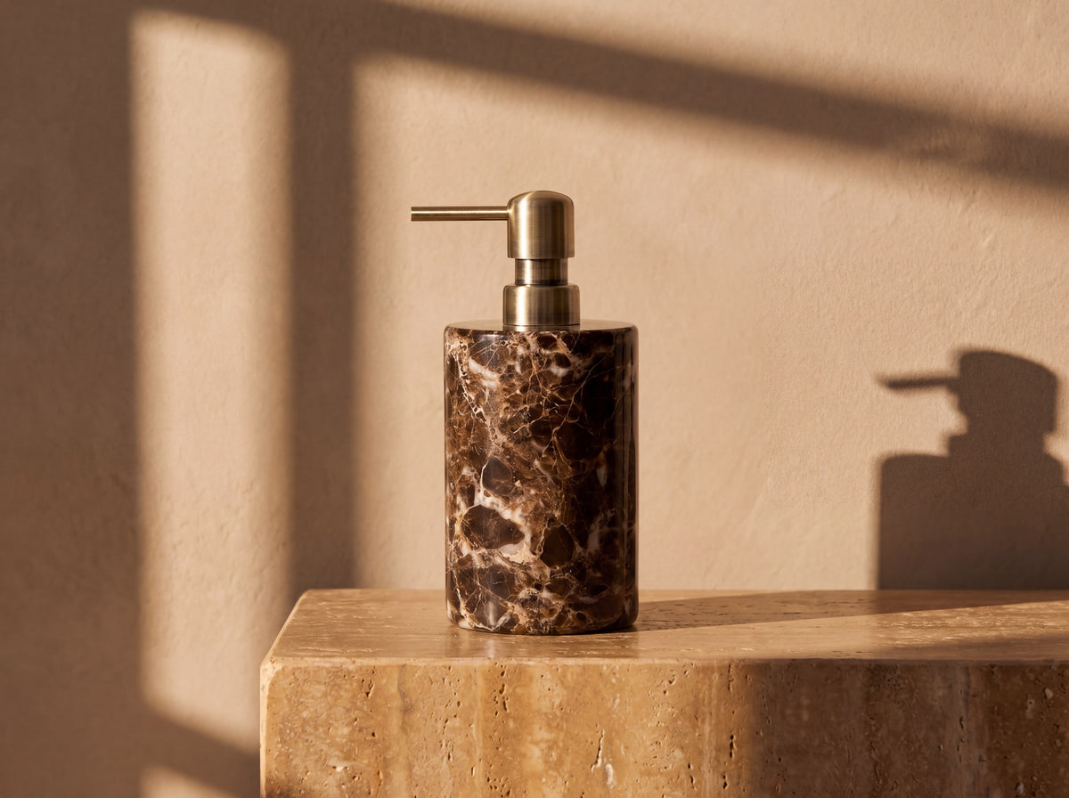 Cylindrical dark Emperador marble soap dispenser with bright white veining and an antique brass pump, lit by warm afternoon sun.