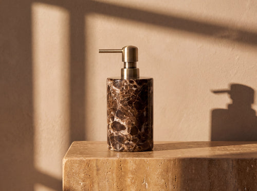 Cylindrical dark Emperador marble soap dispenser with bright white veining and an antique brass pump, lit by warm afternoon sun.