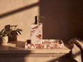 Cream marble soap dispenser with antique brass pump standing on a matching marble tray beside a carved marble soap dish, on a travertine ledge in low golden light.