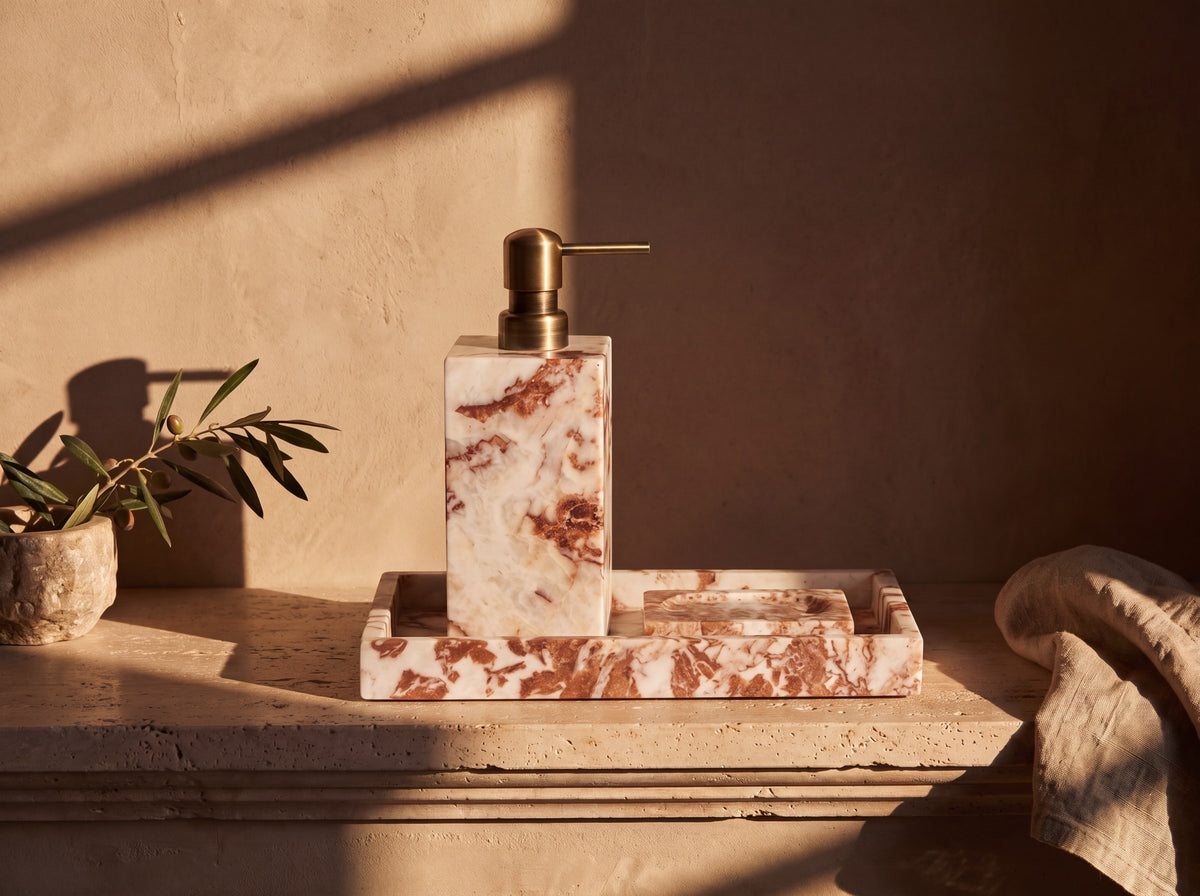 Cream marble soap dispenser with antique brass pump standing on a matching marble tray beside a carved marble soap dish, on a travertine ledge in low golden light.