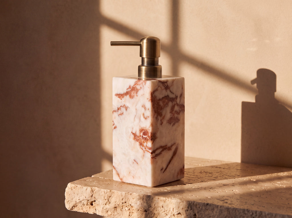 Square column natural marble soap dispenser with rust veining and an antique brass pump, lit by warm afternoon sun on a travertine surface.