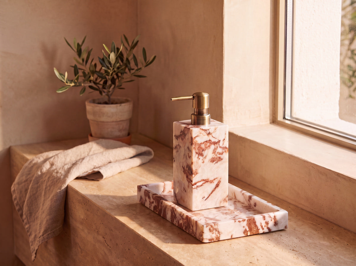 Marble soap dispenser with antique brass pump standing on its matching marble tray on a sunlit travertine window ledge, beside a linen towel and a potted olive plant.