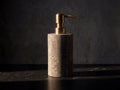 Gray marble soap dispenser with brushed brass pump on a black marble slab, lit by a single warm beam against a charcoal plaster wall.