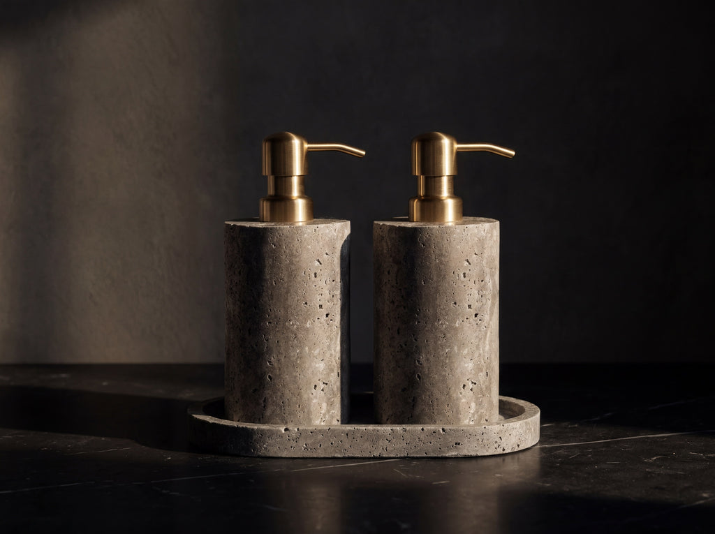 Bathroom set of two gray travertine stone soap dispensers with brass pumps standing on a matching oval stone tray on dark marble.
