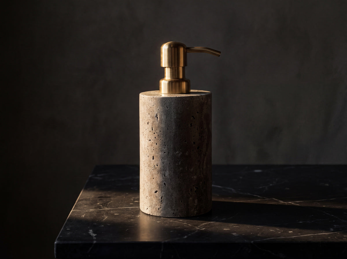 Three-quarter view of a cylindrical gray marble soap dispenser with brass pump against a dark stone backdrop.
