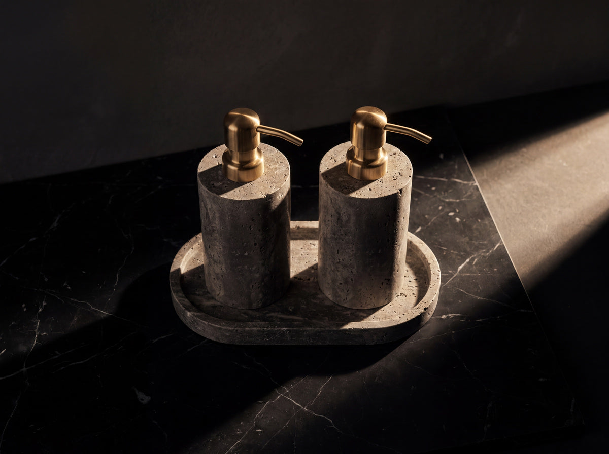 Overhead view of two gray stone soap dispensers on an oval travertine tray, arranged on honed black marble.