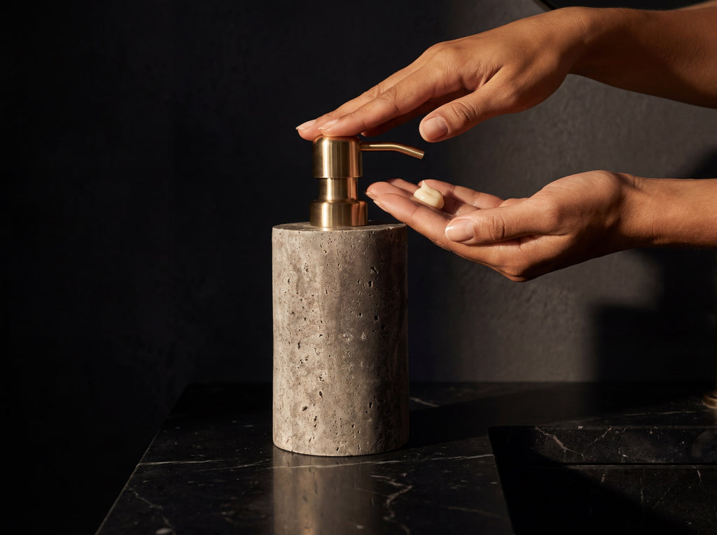 Woman's hand pressing the brass pump of a gray marble soap dispenser, dispensing soap onto her palm at a black marble vanity.