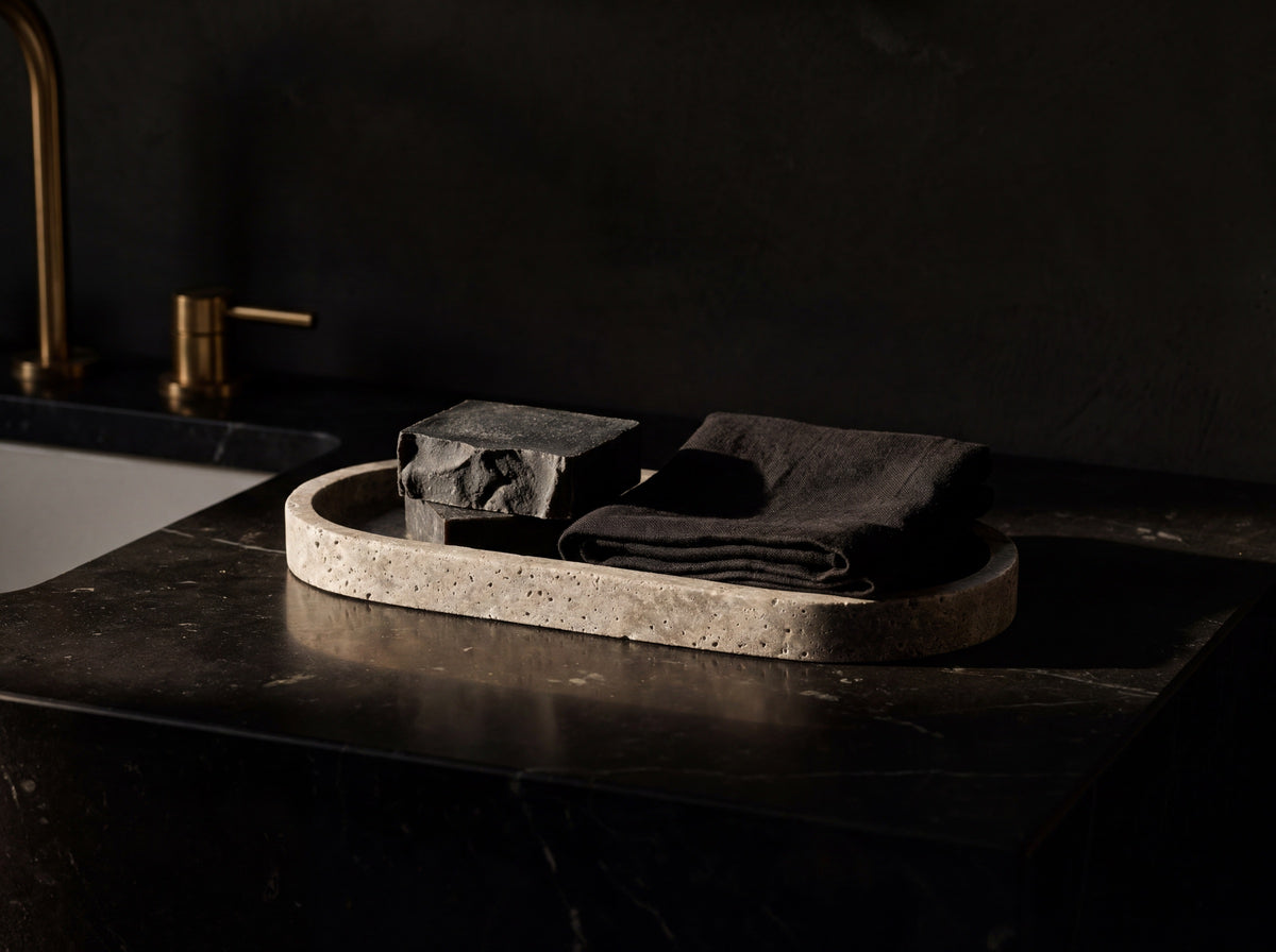 Oval gray travertine tray used as a vanity catch-all holding a charcoal soap bar and folded black linen on dark marble.