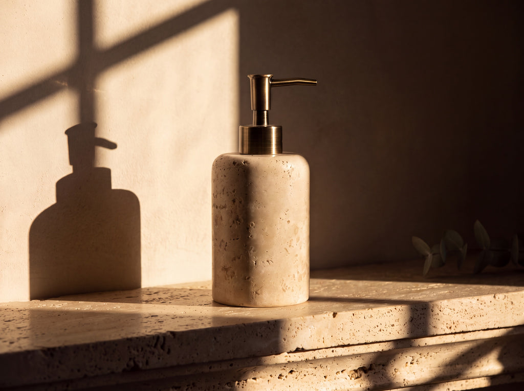 Single travertine stone soap dispenser with antique brass pump on a travertine ledge against warm plaster