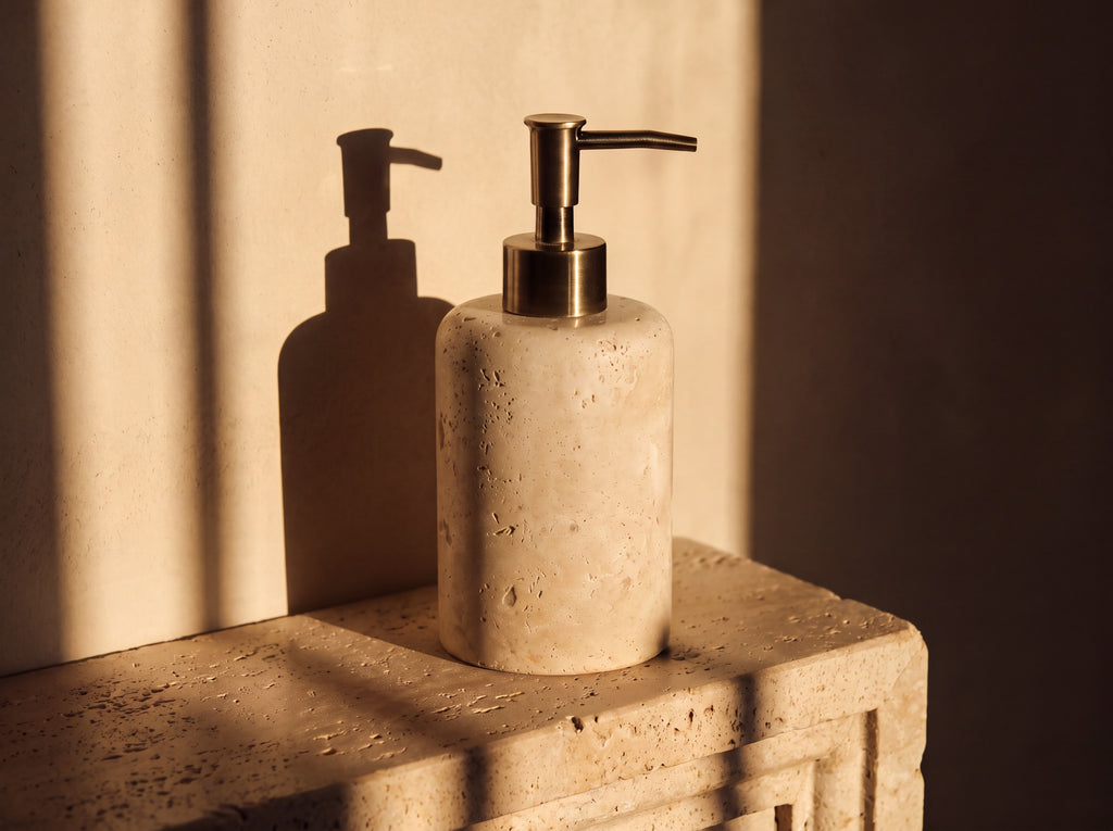 Travertine soap dispenser seen from a three-quarter angle showing the curve of the stone cylinder and brass lever