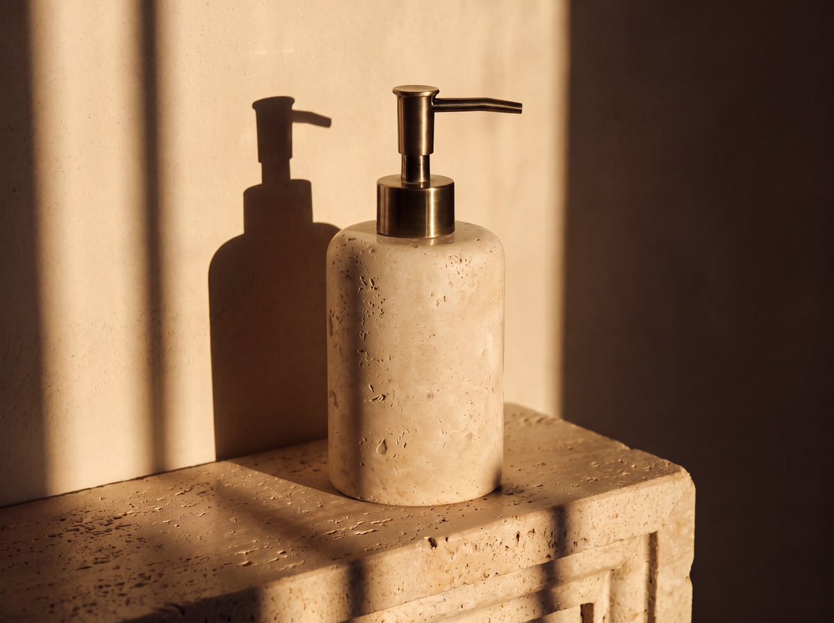 Travertine soap dispenser seen from a three-quarter angle showing the curve of the stone cylinder and brass lever