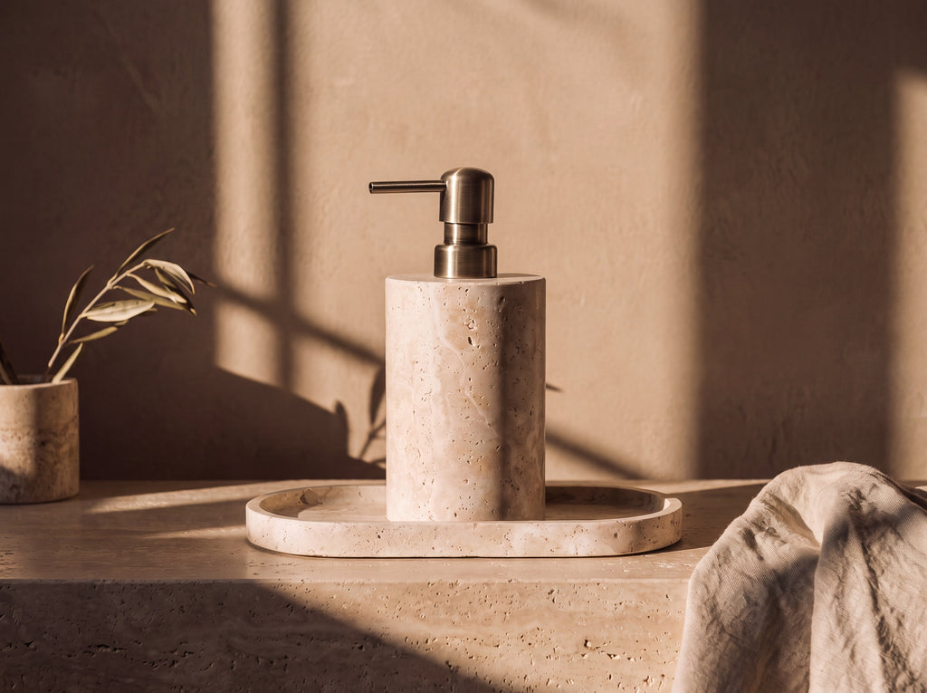 Round cream travertine soap dispenser with a brushed brass pump standing on a matching oval travertine tray on a stone ledge in low golden light.