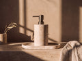 Round cream travertine soap dispenser with a brushed brass pump standing on a matching oval travertine tray on a stone ledge in low golden light.