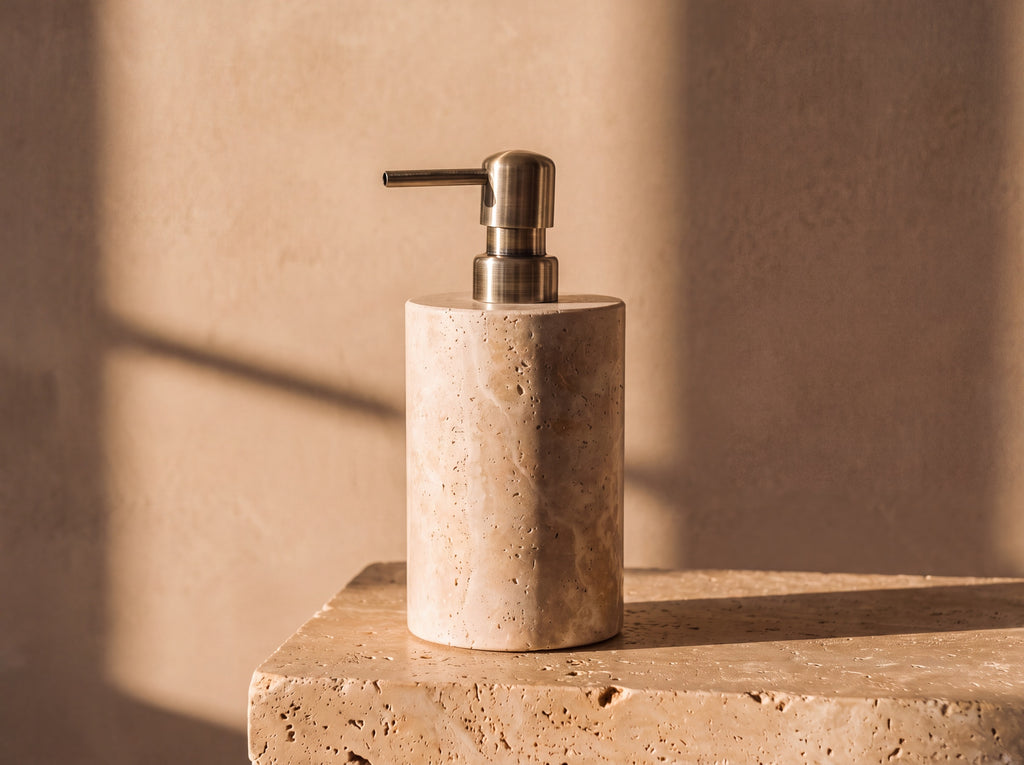 Cylindrical natural travertine soap dispenser with visible stone pores and a brushed antique brass pump, lit by warm afternoon sun.