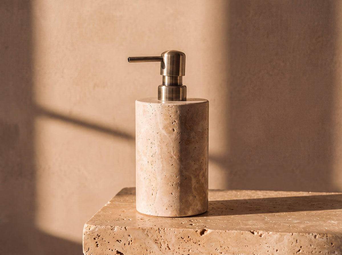 Cylindrical natural travertine soap dispenser with visible stone pores and a brushed antique brass pump, lit by warm afternoon sun.