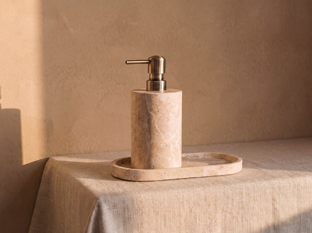 Travertine soap dispenser standing on an oval travertine tray with a raised lip, both cut from the same cream porous stone.