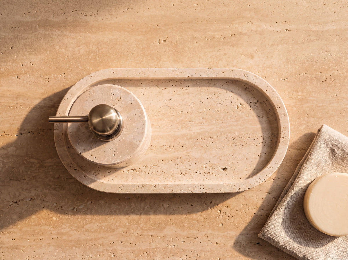 Overhead view of an oval travertine tray holding a round travertine soap dispenser, with a linen cloth and cream soap bar alongside.