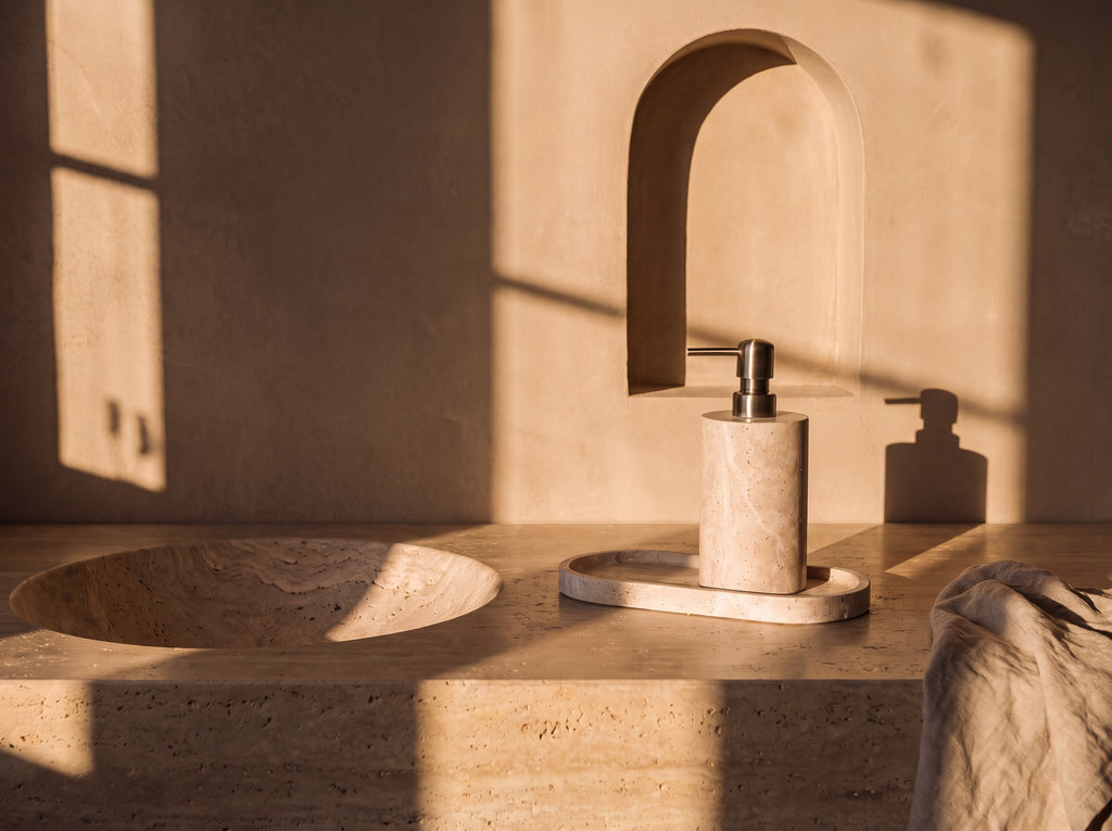 Travertine soap dispenser and oval tray styled beside a carved stone basin on a travertine bathroom vanity.