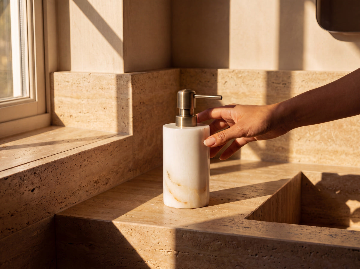 A woman's tan hand resting on the gold pump of a white jade soap dispenser on a sunlit bathroom vanity.