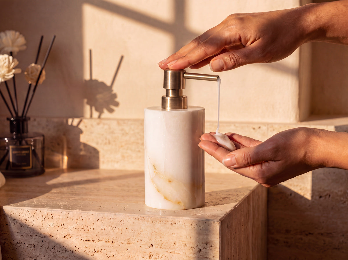 A woman presses the gold pump of a white jade soap dispenser, dispensing a stream of creamy white soap into her cupped hand in warm light.