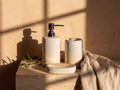 Reactive speckled glaze ceramic soap dispenser, holder and oval soap dish grouped on a travertine ledge in warm sunlight.