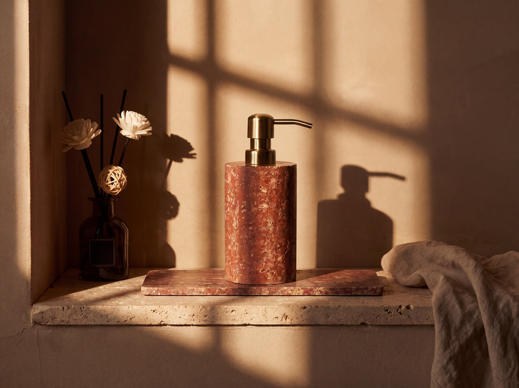 Red travertine soap dispenser with brushed brass pump on a matching stone tray in a sunlit plaster alcove with a reed diffuser and linen cloth