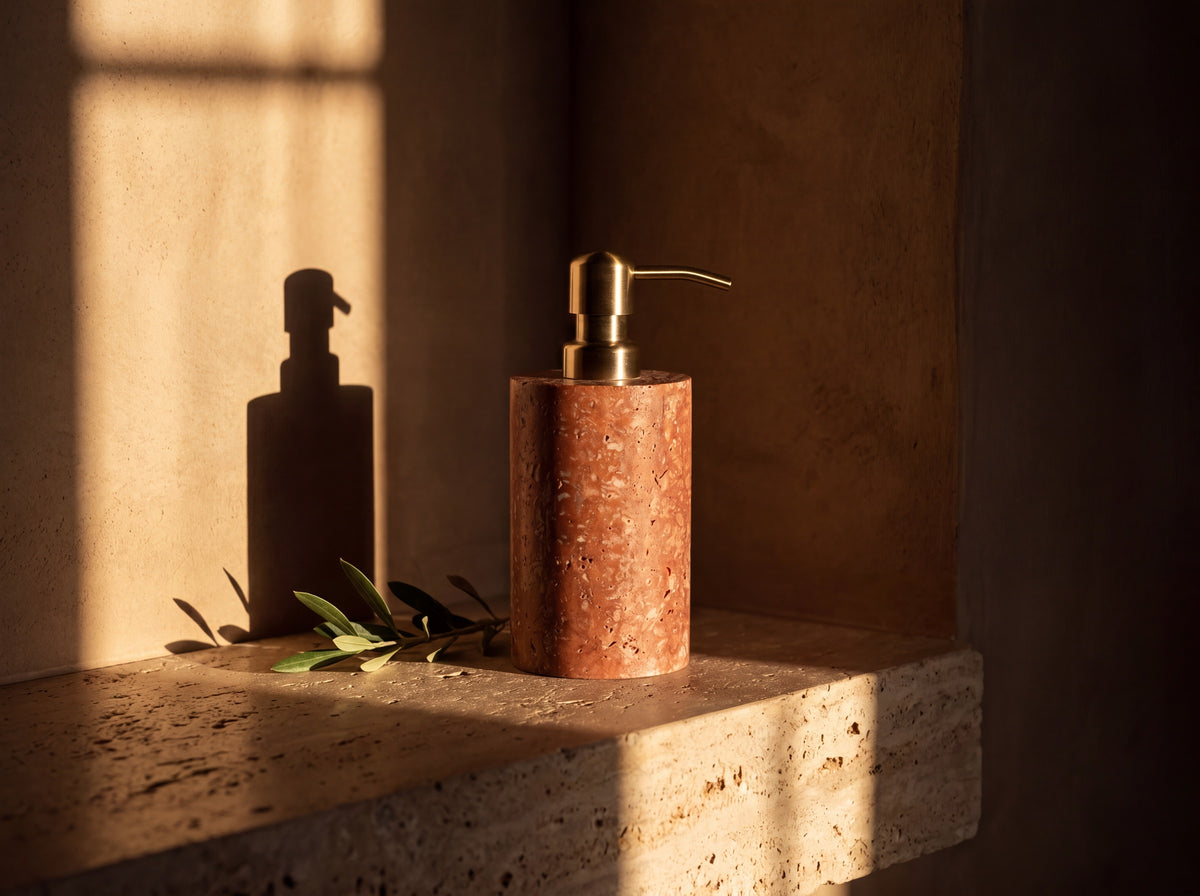 Cylindrical red travertine soap dispenser with brass pump standing on a travertine ledge in low golden light with an olive stem beside it