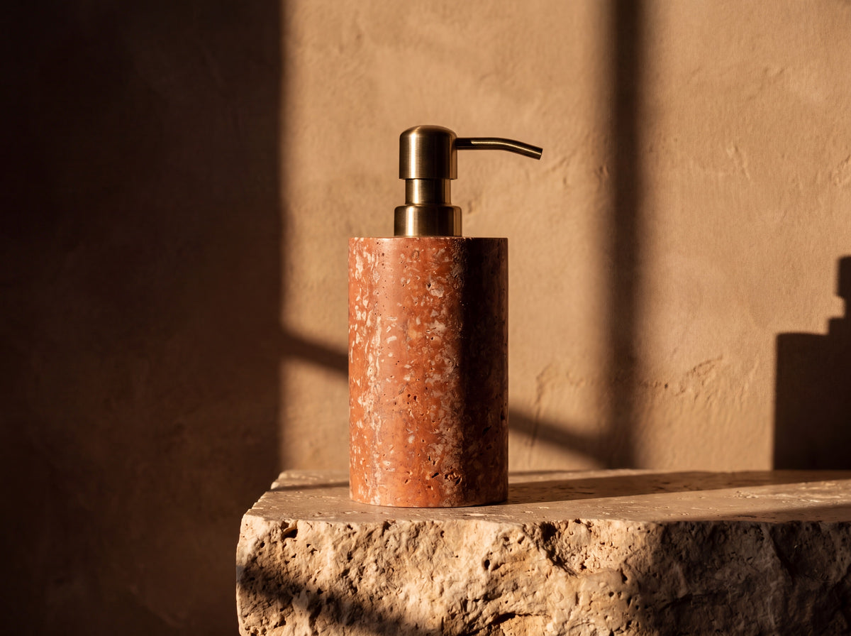 Front view of a red travertine stone soap dispenser with brushed brass pump on a stone ledge against a warm plaster wall