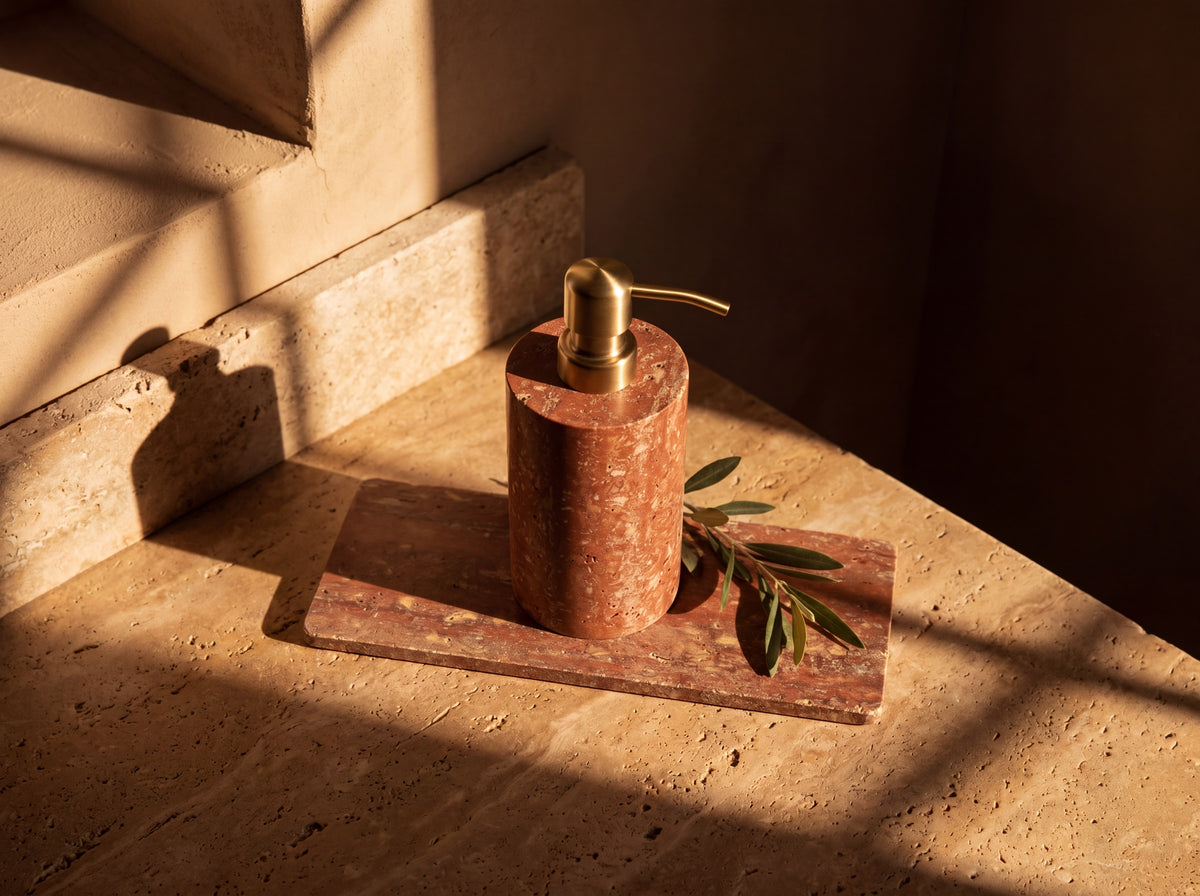 Top-down view of a red travertine soap dispenser standing on its rectangular stone tray with an olive sprig alongside