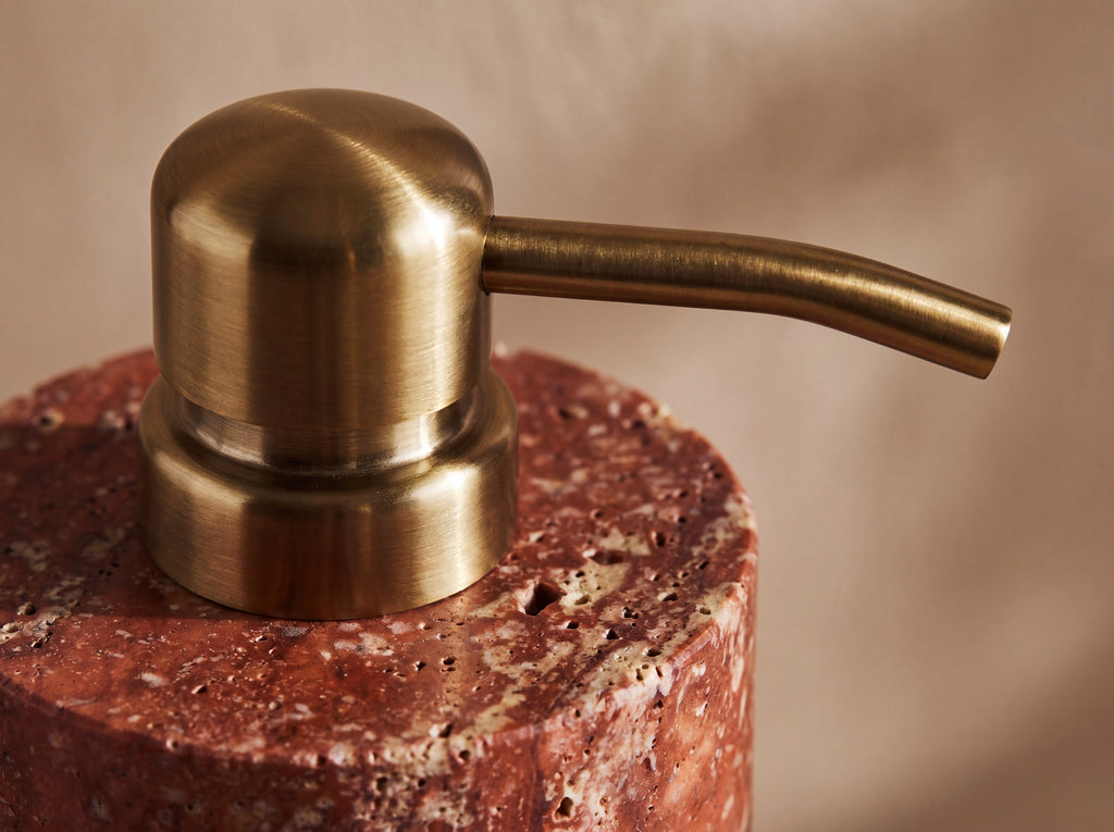 Close-up of the brushed brass pump where it meets the red travertine shoulder of the stone dispenser