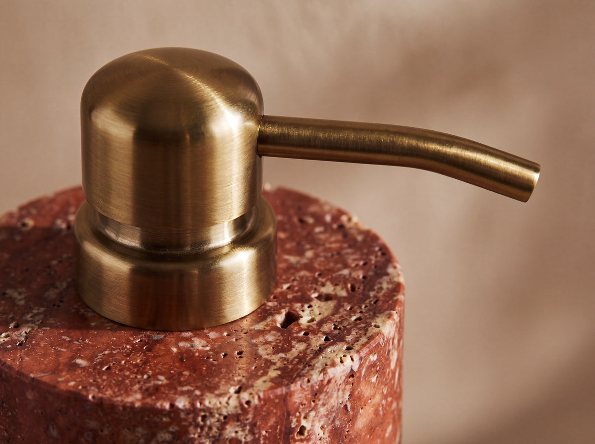Close-up of the brushed brass pump where it meets the red travertine shoulder of the stone dispenser